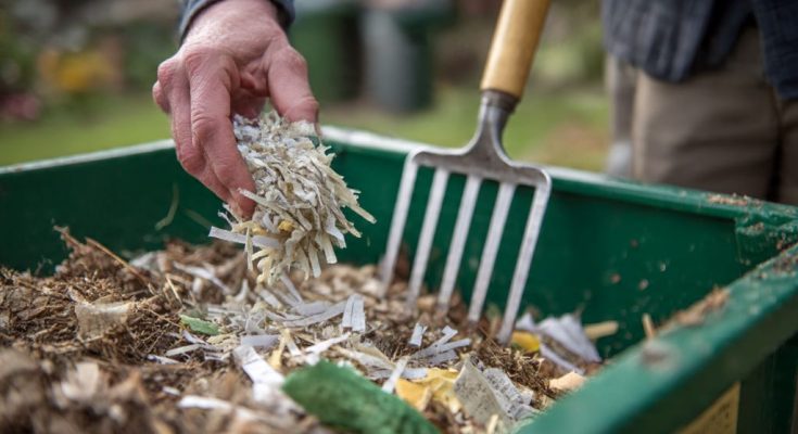 Person adding shredded paper to outdoor compost bin with garden fork and green waste materials visible