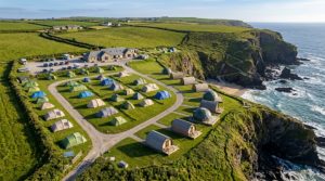 An aerial view of a coastal camping site in Cornwall with pitched tents and glamping pods near the cliff edge, showing prime UK camping land investment potential